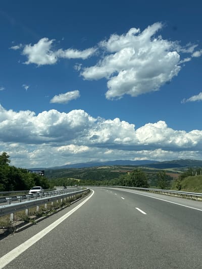 This is a vertical, medium-shot, eye-level photograph of a highway in the city of Blatino, Bulgaria. The highway curves to the right and dominates the foreground, with a white line marking the right edge of the travel lane. A silver guardrail runs along the left side of the highway, with vegetation growing alongside it. In the distance, rolling hills covered in green trees and fields stretch towards a hazy mountain range. The sky is a vibrant blue, filled with large, puffy white cumulus clouds. A few cars are visible on the highway, including a white car on the left and a dark car further ahead. A billboard is visible in the distance on the left, featuring text and graphics. The scene suggests a sunny day with good visibility.