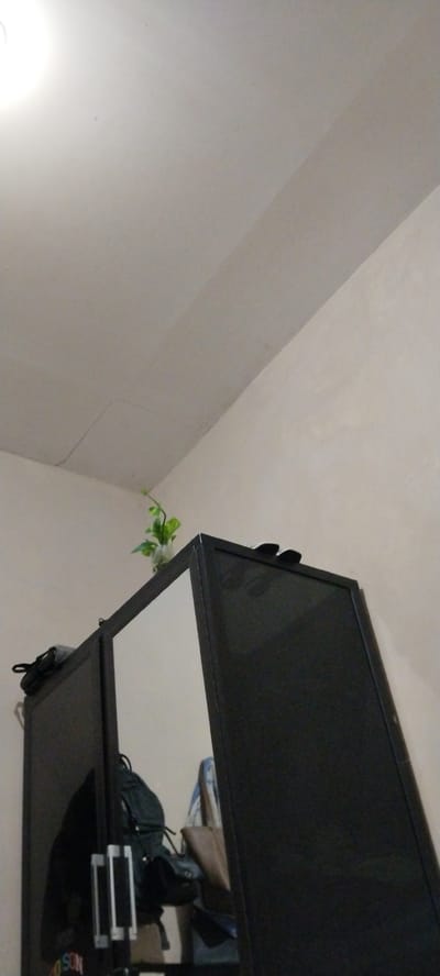 The media file shows the top of a black wardrobe with mirrored doors. On top of the wardrobe, to the left, is a small potted plant with green leaves in a clear vase. To the right of the plant are two dark-colored flip-flops. The wardrobe is situated in a room with cream-colored walls and a textured ceiling. A faint crack is visible in the ceiling above the wardrobe. Reflected in the mirrored doors of the wardrobe are various items, including a dark backpack, a lighter-colored bag, and possibly some clothing. The lighting suggests it might be daytime or indoor lighting is on. The scene appears to be in a domestic setting, likely a bedroom or storage area. No people are visible in the media. The location context is Duman, Indonesia. No visible text is present in the primary image, though there is a suggestion of text on an item reflected in the wardrobe mirror.