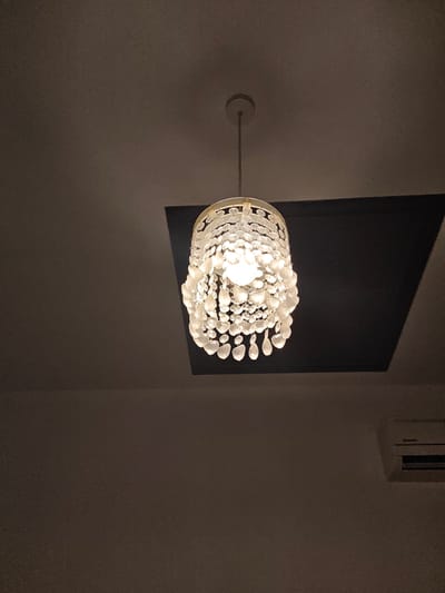 A close-up, low-angle shot focuses on a decorative chandelier hanging from a dark ceiling in a room. The chandelier, with its intricate design of clear, teardrop-shaped ornaments, emits a bright light. Below the chandelier, a dark, rectangular panel is embedded in the ceiling. To the right, a white air conditioning unit is mounted on a plain, dark wall. The lighting in the room is dim, with the chandelier being the primary light source.