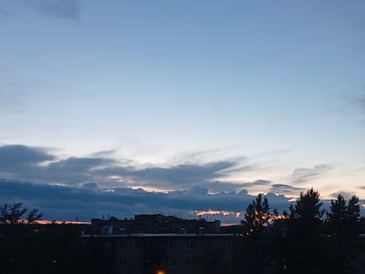 This image depicts a twilight scene over a residential area, likely Kyshtym, Russia. The sky is a gradient of pale blue at the top, transitioning to a lighter hue near the horizon, indicating either sunrise or sunset. Wispy, dark clouds are scattered across the lower portion of the sky, with some illuminated by a faint orange glow from the setting or rising sun, suggesting a transitionary weather pattern.
In the foreground, the silhouettes of trees and buildings are visible. The buildings appear to be apartment blocks, common in Russian residential areas, with visible windows and antenna structures on the rooftops. The overall mood is calm and serene, characteristic of dusk or dawn. The lighting is dim, with the last vestiges of daylight illuminating the sky and casting the foreground elements into shadow. No people are discernible in the image. The image captures a quiet moment in an urban or semi-urban landscape at a time of day marked by subtle color shifts and diffused light.