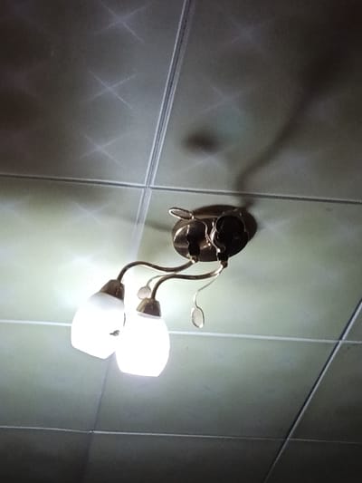 A three-arm ceiling light fixture with frosted glass shades hangs from a tiled ceiling. The light is on, casting a warm glow and creating star-like reflections on the ceiling tiles. A shadow of the fixture and possibly a person is visible on the ceiling. The ceiling appears to be made of square tiles with visible grout lines. The overall lighting suggests it might be evening or indoors with the main light source being the fixture itself. The location is identified as Luhansk, Ukraine, but no specific environmental details are discernible from the image. No text is visible.