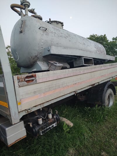 This media file captures an angled view of a large, industrial trailer with a cylindrical tank, parked on a grassy area in Kochmar, Bulgaria. The trailer appears to be made of metal and wood, with visible structural elements and a textured surface. The tank, predominantly light grey, shows signs of wear and peeling paint, suggesting it might be old or in need of maintenance.
Prominent features on the trailer include various pipes and valves attached to the tank, as well as a large tire and suspension system visible near the rear. A section of the trailer's bed is covered with wooden planks, bordered by what appear to be metal strips. The surrounding environment is characterized by lush green grass and foliage, indicating an outdoor setting. A hint of a bright sky suggests it is daytime, possibly late afternoon given the lighting.
The overall impression is one of a stationary, possibly retired or awaiting repair, piece of industrial equipment situated in a natural, unpaved location. No people are visible in the frame, and no specific activity or event is depicted. The image focuses entirely on the inanimate objects and their immediate surroundings, conveying a sense of stillness and perhaps a touch of industrial decay within a rural or semi-rural setting. There is no visible text in the image.