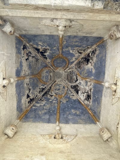 The image shows a close-up, low-angle view of an ornate, frescoed ceiling inside the atrium of the San Andrés Apóstol church in San Andrés Calpan, Mexico. The ceiling is a vaulted structure with intricate stucco work and faded blue paint.

The stucco features eight radial beams, each adorned with gold-colored rope-like detailing and terminating in carved angel-like figures at the corners of the ceiling. These figures have wings and stylized faces. In the center of the ceiling, a circular stucco medallion contains an embossed religious symbol, possibly "IHS" for Jesus. The blue paint on the ceiling is peeling and distressed, revealing darker plaster beneath. The overall impression is one of aged grandeur and historical significance.