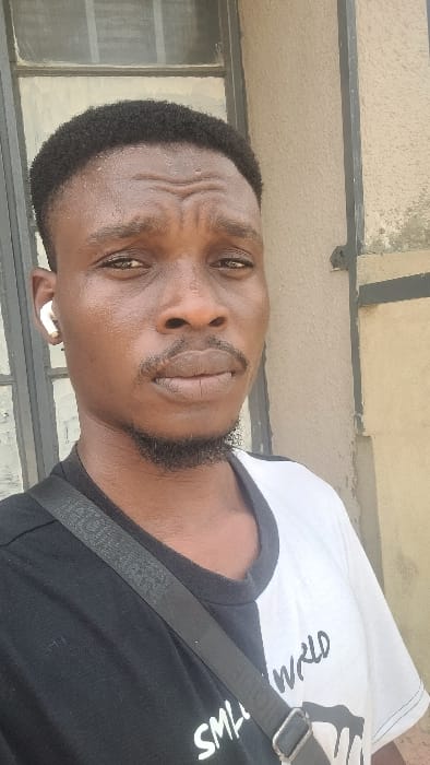 The scene depicts a sole male individual, approximately 20-35 years old, of African descent, in a close-up portrait, presumably taken in Badiko, Nigeria. The subject possesses short, dark, coiled hair and a dark mustache and goatee. His facial skin, particularly the forehead, appears damp with perspiration, showing horizontal stress lines. He wears a t-shirt bisected vertically: black on the left, white on the right, with "SMALL WORLD" partially discernible in black lettering on the white portion. A black fabric strap, consistent with a messenger bag or sling, diagonally traverses his left shoulder and chest. A white wireless earbud is inserted into his left ear. The background consists of a window with dark framing and an opaque light-colored pane on the viewer's left, and a textured, tan-colored exterior wall exhibiting minor wear to the viewer's right. The subject is looking directly at the camera.