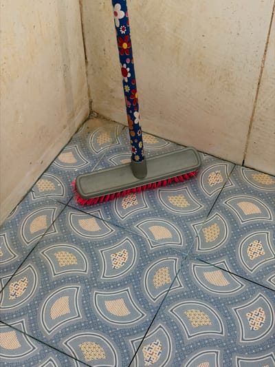 The image shows a close-up view of a broom with a colorful floral-patterned handle and a grey head with pink bristles, positioned on a tiled floor. The floor has a repeating pattern of blue and white semi-circular motifs containing smaller textured shapes. The broom is in a corner where two walls meet. The walls are a light beige color and appear to be tiled or paneled, with some visible grime and discoloration, particularly in the corner. The overall scene suggests a cleaning activity or the presence of cleaning tools in a somewhat aged or neglected environment, possibly a bathroom or utility area. The location is stated to be Tupah, Indonesia.