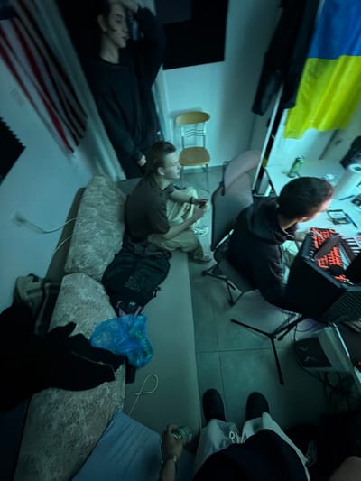 This image captures an indoor scene within a room located in Migdal HaEmeq, Israel, illuminated by a cool, bluish-green ambient light. Four individuals are present.
In the upper-left, a person with long dark hair, wearing a dark long-sleeved shirt, stands partially obscured by shadow, looking towards the right with a hand near their head. Below them, a person with medium-length light brown hair, dressed in a dark t-shirt and light-colored trousers, sits on a light-colored sofa with patterned cushions. This individual holds a small dark electronic device and looks towards the right. A black backpack rests on the sofa beside them.
To the right, a person in a dark hooded sweatshirt is seated at a white desk, actively interacting with a laptop featuring a glowing red keyboard. A green beverage can and various small objects are arranged on the desk. This individual is supported by a dark office chair.
The lower portion of the image shows the legs and lower torso of a fourth person, clad in light-colored pants and dark shoes. A left hand, wearing a watch, holds a green can.
The room's environment includes white walls. A red and white striped flag is partially visible on the left wall, and a yellow and blue flag is draped on the right wall. A black acoustic foam panel is affixed to the left wall. A light-brown wooden chair with a metal frame is positioned near the center. The floor is composed of light-colored tiles. A white power outlet with a connected white cable is visible on the left wall near the sofa, and a black tripod stand is located near the desk. A blue plastic bag lies on a sofa cushion in the foreground.