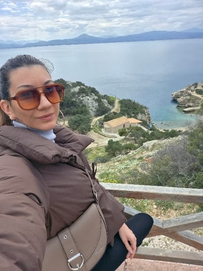 The image depicts a female subject in a selfie-style composition from an elevated viewpoint in the Municipal Unit of Loutraki - Perachora, Greece. She is positioned in the left foreground, wearing dark brown-tinted sunglasses, a light blue turtleneck, and a brown quilted puffer jacket. A beige shoulder bag with a silver D-ring buckle is draped across her chest. Her dark hair is pulled back.
Below her, a rocky, vegetated hillside descends steeply towards a large body of water. A rustic wooden railing is visible in the lower right foreground. In the midground, a small, light-colored building with a terracotta roof stands on a promontory, accessible by a winding dirt path. To the right of the building, a secluded cove with clear, light blue water and a small beach is visible, bordered by rocky cliffs; a small Greek flag is mounted near the building. The background features a vast expanse of calm, deep blue water leading to a distant mountainous coastline under a predominantly cloudy, grey sky.