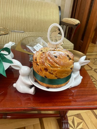 A tilted medium shot depicts an Easter kulich on a white plate, adorned with a green ribbon and white ceramic bunnies, placed on a polished wooden surface. The kulich is garnished with raisins and dusted with powdered sugar. An Easter egg decoration on a stick is inserted into the kulich. Part of a gold-rimmed glass display case can be seen on the left. The background features a patterned wall and furniture. The location is likely a home in Akhalkalaki, Georgia, based on the kulich, a traditional Easter bread of Orthodox Christian cultures.