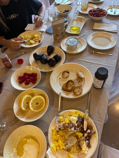 A table is laden with various plates of food, suggesting a shared meal. Plates contain scrambled eggs, prosciutto, salad, toast, avocados, raspberries, and lemon slices. A bowl of strawberries, a jar of jam, a tin of sardines, and a dish of cheese are also present. A person is eating from a plate of scrambled eggs. The spread suggests a brunch or breakfast setting.