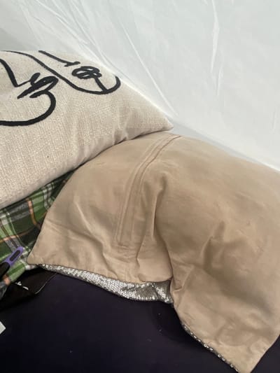 The image shows a pile of soft furnishings, including a light beige pillow with a quilted texture and black abstract line art, and a larger, smooth beige pillow. A dark navy fabric is visible on the right, possibly a couch or a blanket. A strip of silver sequins runs along the edge of the beige pillow. Peeking out from underneath is a green and white plaid fabric. The overall impression is one of domestic comfort, though the close-up framing and the plastic sheeting on the left suggest it might be in storage or being packed. The lighting is bright and diffuse, indicating it's daytime. There is no visible text.