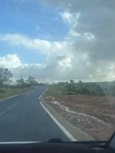The image depicts a rural road scene in Oumazza, Morocco, viewed from inside a moving vehicle. A two-lane asphalt road, dark grey and appearing damp, curves gently left then right, receding into the mid-ground. It features a solid white line marking the right edge and a dashed white line in its center. To the immediate right of the road, a narrow grey shoulder borders a muddy, reddish-brown dirt strip exhibiting standing water and rivulets, indicative of recent precipitation. Beyond this, a gradual uphill slope is covered with green grass, scattered bushes, and small trees.
On the left side of the road, similar green vegetation and numerous trees line the route. Multiple utility poles, dark in color, are visible on both sides of the road in the mid-distance. The sky is partly cloudy; the upper left quadrant features clear blue sky with bright white cumulus clouds, while the upper right and central portions are dominated by larger, darker grey stratocumulus clouds. No individuals or animals are present.