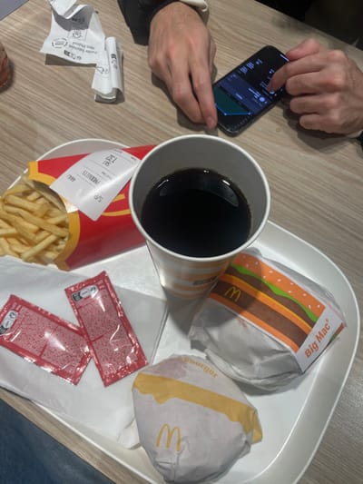 An aerial shot reveals a person using a smartphone next to a McDonald's meal on a tray. The meal includes a Big Mac, another burger, fries in a red McDonald's container, two packets of ketchup, and a large cup filled with a dark beverage. Crumpled receipts lie on the table beside the tray. The receipts display numbers and words, possibly related to the McDonald's order.