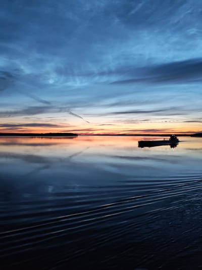The image depicts a serene twilight scene over a body of water, possibly a large lake or calm sea, with a cargo ship silhouetted against the horizon. The sky is a gradient of deep blue at the top, transitioning to lighter blues and oranges near the horizon where the sun has recently set, casting a warm glow. Wispy clouds streak across the sky, some catching the last rays of sunlight. The water's surface is calm, reflecting the colors of the sky and creating a mirror-like effect. A cargo ship is visible in the mid-ground, moving from right to left, leaving a wake of rippling water behind it. The far shore is lined with a dark silhouette of trees, suggesting a natural landscape rather than an urban one. The overall impression is one of tranquility and the vastness of nature at dusk. There is no discernible connection to the city of Malinukha, Russia in the image.