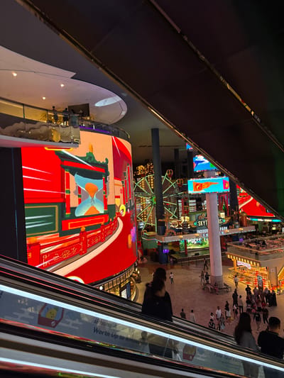Captured from an elevated vantage point, the image presents a vibrant scene at Genting Highlands, Malaysia. The perspective is likely from an escalator, offering a view of the complex's bustling interior. The focus is drawn towards a large LED screen showcasing colorful, animated graphics with a distinct Asian aesthetic.
Below this screen, a Ferris wheel decorated with lights adds to the lively atmosphere. Signage for "SkyTropolis Indoor Theme Park" and "Swensen's" is visible, indicating the entertainment and dining options available.
Several people are milling about in the area, contributing to the overall activity. The presence of architectural elements such as pillars and railings further contextualizes the scene within an enclosed, multi-level structure.