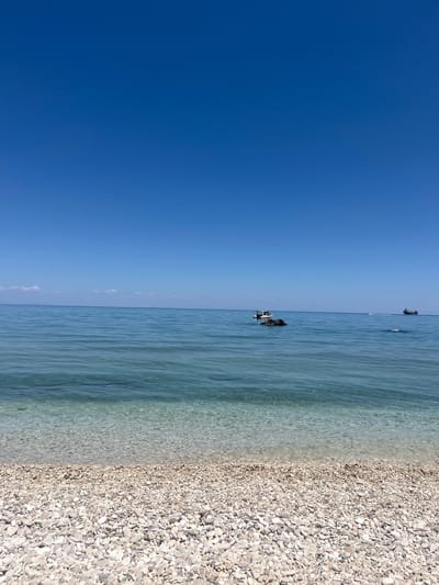 The image depicts a serene seascape with a clear blue sky meeting the calm, turquoise waters of the sea. In the foreground, a pebble beach is visible, with the water gently lapping at its edge. The shallow water near the shore is transparent, revealing the smooth pebbles beneath. Further out in the water, several boats are present. A small white boat with an orange canopy is anchored near a rock formation. In the distance, a larger dark vessel, possibly a cargo ship, is sailing on the horizon, accompanied by a smaller white boat. The overall scene suggests a peaceful day at the coast in the Elaties Municipal Unit, Greece, characterized by its natural beauty and tranquil atmosphere.