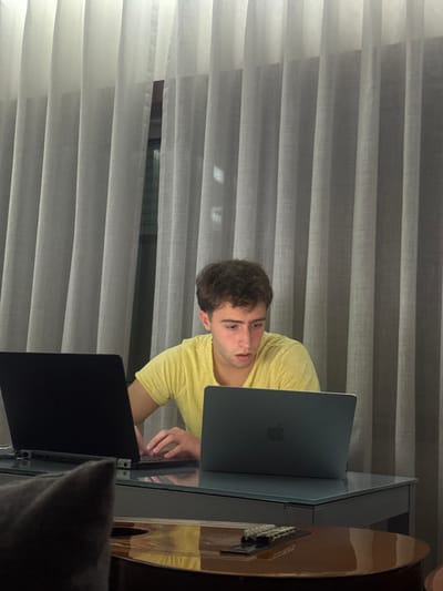 A young man with short curly hair and a yellow t-shirt is seated at a glass table, operating a dark-colored laptop. Another laptop sits in the foreground, partially obstructed by a gray blanket or pillow. A guitar rests on the table to the right of the man. Vertical sheer curtains cover the background, allowing some light to filter through. A dark horizontal line, likely a window frame, is visible behind the curtains.