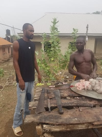 The image depicts two men standing near a wooden table, with one man in the foreground and one man in the background. The foreground man is wearing blue jeans and a black sleeveless t-shirt. He is standing with his right leg slightly bent.  The background man is bare-chested and is holding a knife with his right hand. The background man is preparing a cut of meat on a large metal pan situated on top of the wooden table. The meat is unidentifiable, but it appears to be raw and possibly offal. Three long, black knives are also present on the table next to the metal pan. There are other unidentified objects on the table. The image appears to have been taken outside in a residential area. The location is Pati Ridge, Nigeria. The coordinates of the location are 7.81 latitude and 6.704 longitude.  There are buildings in the background. The area appears to be relatively clean. The sky is overcast and gray.  The overall scene appears to be one of casual butchering and preparation. The men appear to be working together, though there is no evidence of conversation or interaction. The tone of the image is matter-of-fact.