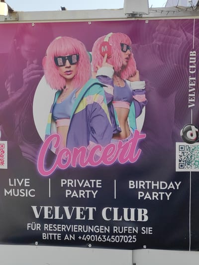 This is a photograph of a poster displayed outdoors. The poster advertises a "Velvet Club" and features an image of two women with pink wigs and futuristic sunglasses. The women are wearing colorful outfits and headphones. The text on the poster includes "Concert", "Live Music", "Private Party", and "Birthday Party". Below this, it states "Velvet Club" and provides contact information for reservations in German: "FÜR RESERVIERUNGEN RUFEN SIE BITTE AN +4901634507025". There is also a TikTok logo and two QR codes. The poster is mounted on a white surface, possibly a building wall, with visible wires and lighting fixtures along the top edge. The overall impression is a promotional advertisement for a nightlife venue. The city of Brühl, Germany is not explicitly visible or referenced in the image, only the club's name is provided.