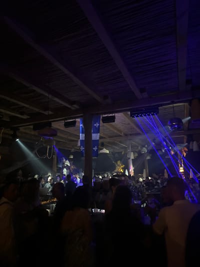 The image captures a crowded scene inside a venue, likely a nightclub or bar, during nighttime. The ceiling is constructed from thatched material supported by wooden beams, and is illuminated by blue and white lights, including visible laser beams cutting across the space. Several hanging light fixtures are present. On the right side, a disco ball is visible. Two blue banners with a white fleur-de-lis symbol hang from the ceiling. A crowd of people are visible in the foreground and background, many standing and some seated at tables, appearing to be socializing and enjoying the atmosphere. The overall ambiance suggests a lively entertainment setting. The location is identified as Asprovalta, Greece.