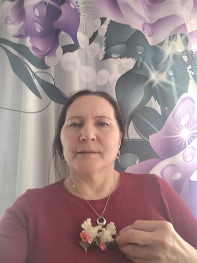 A close-up, eye-level portrait features a middle-aged woman with light skin and dark hair, looking directly at the camera with a neutral expression. She is wearing a maroon or dark red long-sleeved top. Around her neck is a silver-colored chain with a prominent pendant. The pendant appears to be crafted from a light-colored material, possibly bone or resin, depicting two bird-like figures, likely doves, intertwined with small pink/red flowers and green leaves. Her right hand is gently holding or touching the pendant.
The background consists of a brightly lit, patterned curtain, possibly a shower curtain, featuring large, stylized purple floral motifs and dark green leaves or stems. Interspersed within this pattern are numerous white, translucent, circular shapes resembling bubbles or raindrops. Several areas of the background display bright, starburst-like reflections, suggesting a strong light source or glossy material. The far-left side of the background shows a distinct pattern of vertical white and light blue stripes. The scene is set indoors, within what appears to be a domestic interior in Сусады-Эбалак, Russia.