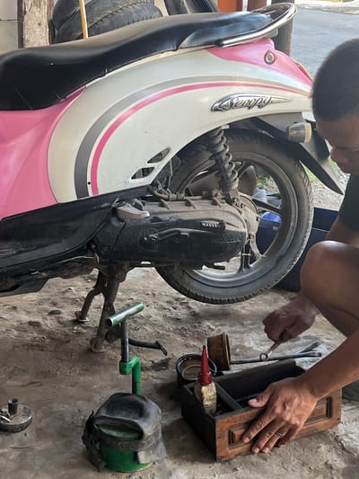 A man is repairing a pink and white Honda Scoopy motorcycle in Paweden, Indonesia. The motorcycle's rear wheel is visible, as is the engine. The man is using tools from a wooden toolbox. A green device and other motorcycle parts are scattered on the ground. The scene is set outdoors on a concrete surface.