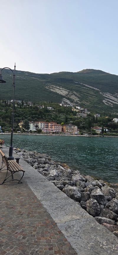 The image depicts a scenic view of the lakefront in Torbole, Italy. In the foreground, a stone embankment lines the water, with a paved walkway made of reddish-brown cobblestones. A weathered wooden bench with metal legs is placed on the walkway, next to a black lamppost. The lake, a deep blue-green, extends towards the background, where a lush green mountainside rises. Dotted along the hillside are various buildings, some appearing to be hotels or residences, with colorful facades. The sky above is a clear, pale blue, with a hint of haze. The overall impression is one of tranquility and natural beauty.