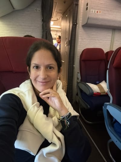A woman is seated in an airplane, likely in the economy cabin based on the seat spacing. She is wearing a white sweatshirt over a black shirt and has bracelets on her left wrist. She appears to be smiling and looking at the camera. Behind her are red airplane seats and the aisle. In the distance, a flight attendant wearing a blue shirt is visible. The scene is likely inside an aircraft, but nothing in the image specifically identifies the location as Pudahuel, Chile. The location is unknown from the image.