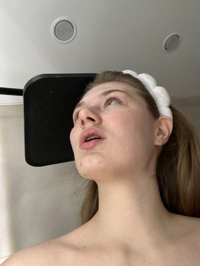 A woman with light skin and blonde hair tied back with a white headband reclines against a black chair. The camera angle is low, shooting upwards at her face. Her mouth is slightly open, and she appears to be looking towards the upper left corner. Two circular ceiling lights are visible in the background, which appears to be a white tiled room, potentially a bathroom or spa.