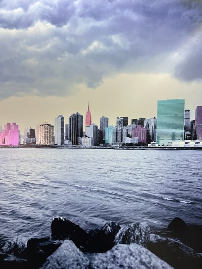 The image depicts a wide shot of a city skyline viewed across a body of water. The skyline is composed of numerous skyscrapers of varying heights and architectural styles. Some buildings are rendered in grayscale, while others are highlighted with vibrant, artificial colors, including pink, teal, and purple. The most prominent colored building is a tall, rectangular teal structure on the right side of the skyline. In the center, a distinctively shaped, pink-hued skyscraper resembling the Empire State Building is visible. In the foreground, the dark, rocky shoreline of the body of water is visible, with waves gently lapping at the shore. The sky above is filled with dramatic, heavy clouds, with a muted yellow and purple gradient towards the horizon, suggesting either dawn or dusk. The overall impression is a stylized, artistic representation of a major metropolitan area, possibly New York City, with a deliberate use of color to emphasize certain structures against a more muted background. The location is not Schoonderbuken, Belgium.