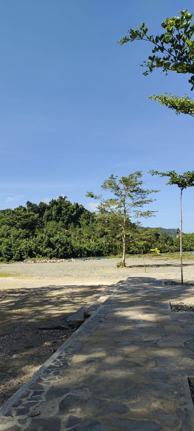 The image shows a paved walkway made of irregular stone slabs leading towards a natural landscape under a clear blue sky. In the foreground, the walkway is illuminated by dappled sunlight casting shadows. Beyond the walkway is a gravelly area, followed by a body of water with a rocky bank. A lone tree stands in the mid-ground, casting a shadow onto the gravel. In the background, a dense, green forest covers a hillside, with hints of palm trees visible. The sky is a vibrant blue with no clouds. The scene appears to be a park or recreational area in Lamseunia, Indonesia, given the lush tropical vegetation and clear weather. There are no people or animals visible in the image.