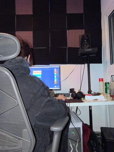 An individual is seated in a grey ergonomic office chair, facing away from the camera, engaging with a computer setup within a room in Migdal HaEmeq, Israel. The person wears a dark, possibly black, denim jacket or heavy hoodie, with short dark hair visible. Their hands are positioned over a keyboard and mouse on a white desk.
The computer monitor displays a Digital Audio Workstation (DAW) software interface featuring multiple colored horizontal tracks. To the right of the monitor, on the desk, a black studio monitor speaker labeled "Alctron" is mounted on a stand. Various items are present on the white desk, including: a brown glass bottle, a green aluminum can, a red aerosol can, a pair of black headphones, and a small black electronic device. Notably, a small pile of green, leafy, granular substance rests on a crumpled piece of white paper or tissue. A white PC tower with illuminated internal components is visible on the floor beneath the desk.
The wall behind the computer setup is covered with sound-absorbing acoustic foam panels, arranged in a checkerboard pattern of dark grey/black and reddish-brown or dark purple sections. A white wall and a window frame are visible on the right side of the image. Multiple black cables are connected to the electronic equipment.