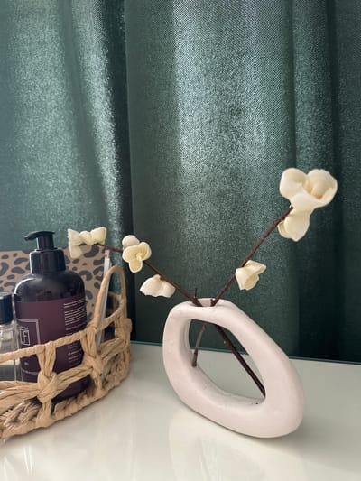 This image depicts a still life scene on a white reflective surface, with a dark green textured curtain in the background. The scene features a woven basket containing a dark purple pump bottle of lotion, partially obscured by the basket's weave. To the right of the basket, a minimalist, pale pink vase with an oval opening holds a delicate branch with small, white artificial flowers. The overall aesthetic suggests a decorative arrangement, likely indoors. There is no indication of the city of Malye Ust'ya, Russia, or any people, actions, or interactions within the image.