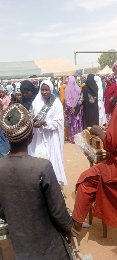 This image captures a crowded outdoor scene in Sani Mai Nagge, Nigeria, under a bright, slightly cloudy daytime sky. The ground is exposed earth.
In the mid-ground, a young adult woman stands centrally, dressed in a white hijab and a long white garment. She wears a black sash diagonally across her chest, bearing white text that includes "2026" and Arabic script. Her mouth is slightly open as if speaking, and she faces towards the left foreground. To her immediate left, another woman in a black hijab and dark top stands beside her.
In the left foreground, a person is viewed from behind, wearing a dark long-sleeved top and a patterned, woven brown and white cap. This individual is positioned to face the woman in white. In the right foreground, a person in a red long-sleeved garment is partially visible, seated, with their hands positioned near a display of numerous rectangular, light-brown blocks. These blocks are stacked in rows on a stand and appear to be individually wrapped in clear plastic.
The background features a dense gathering of people, primarily women, dressed in various colorful hijabs and traditional attire, including purple, red, orange, and white garments. Several large tents, some striped in blue and beige, are erected behind the crowd, suggesting a market or event setting. Distant trees and a metal frame structure are visible in the far background.
