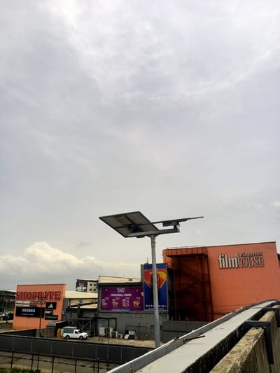 The image shows a commercial area in Eputu Town, Nigeria, under an overcast sky. Visible retail establishments include "SHOPRITE" and "filmhouse cinemas." There is also a billboard advertising a "CIRCLEMALL COMBO." Another billboard features an image of Supergirl. A solar-powered street light is in the foreground, along with a concrete barrier. A white van is parked in the lot below. Other smaller retail logos such as Adidas, Puma, Skechers, and Miniso are visible. An external staircase is attached to the "filmhouse" building. Several parked cars are visible in the background.