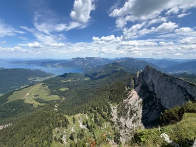 This is a panoramic, high-angle view of a mountainous landscape near Sankt Gilgen, Austria. The scene captures a vast expanse of rolling hills covered in dense green forest, leading to a large, calm lake in the distance. Towering, rugged mountains with sheer rock faces dominate the right side of the frame, casting shadows across the slopes. The sky above is a vibrant blue, dotted with scattered white cumulus clouds. A winding path is visible on a grassy slope in the mid-ground, leading through patches of trees. In the foreground, the edge of a grassy cliff overlooks the expansive vista.