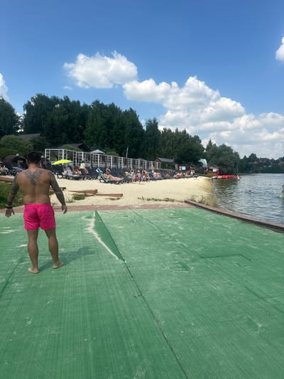 The image depicts a sunny day at a lakeside resort in Dal'nie Prudishchi, Russia. In the foreground, a man with extensive back tattoos and pink shorts stands on a green, textured surface, facing away from the camera. The surface appears to be a walkway or ramp leading towards the water. Behind him, a sandy beach is lined with rows of lounge chairs, many occupied by people sunbathing or relaxing. Further back, there are structures resembling cabanas and buildings, with a dense line of trees forming the backdrop. The lake is visible to the right, with several people in the water and some red pedal boats docked near the shore. The sky is blue with scattered white clouds.
