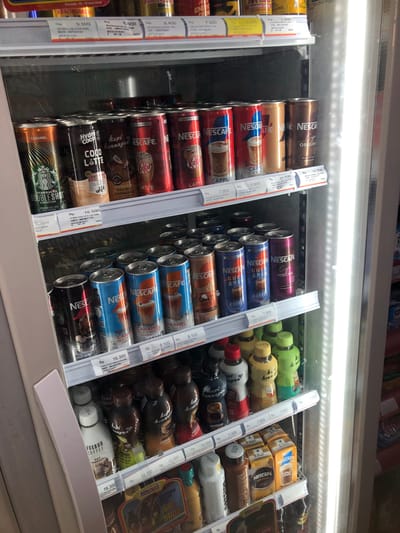 The image shows the inside of a commercial refrigerator, likely in a convenience store or minimarket in Putat, Indonesia, as indicated by the text "Alfamart" visible on a price tag. The refrigerator is stocked with various beverages, primarily canned coffee drinks and bottled coffee drinks.

On the top shelf, there are several rows of canned coffee. Brands visible include Nescafe and Hydro Coco. The cans come in various flavors such as Caramel Macchiato, Cappuccino, and Original. Some cans have red, blue, and brown labels.

The middle shelf also contains canned coffee, with many Nescafe cans. Some are labeled "Ice Blended," "Caramel Macchiato," "Gula Aren," and "Mocha." There are also some other canned drinks with gold and brown labels.

The bottom shelf holds bottled drinks. There are several white bottles that say "FOXBUD COFFEE OAT," and other brown and multi-colored bottles with text that appears to be Indonesian. Some of these bottles are branded "Kopi Kenangan" and "Kopi Susu Alpurat." There are also small yellow boxes of Nescafe Coffee Creamer.

Price tags are visible throughout the shelves, indicating prices in Indonesian Rupiah (Rp). The lighting in the refrigerator is bright and white, likely from LED strips, illuminating the products. The overall scene is neat and organized, suggesting a well-stocked retail environment.