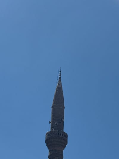 A close-up, low-angle shot captures the upper portion of a dark, conical structure against a clear, bright blue sky. The structure appears to be made of stone or concrete and exhibits a rough, textured surface with visible horizontal banding. It narrows to a pointed tip, adorned with several small, spherical ornaments suspended from thin wires, resembling tiny bells or finials. The larger part of the visible structure features arched openings or windows, hinting at architectural detail. The overall impression is of a minaret or spire. The bright sky suggests it is daytime, likely with good weather conditions. The image is cropped tightly on the object, with the vast expanse of the sky dominating the background, providing no other contextual cues for location beyond the information provided. No text is visible in the image.
