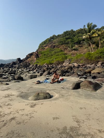 A beach scene shows a man on a towel, positioned near rocks and a cliff covered in green foliage. The sky is clear and blue. A large, round rock embedded in the sand creates a distinct feature on the sandy shore. The orientation of the image is vertical, with the shoreline running along the right side.