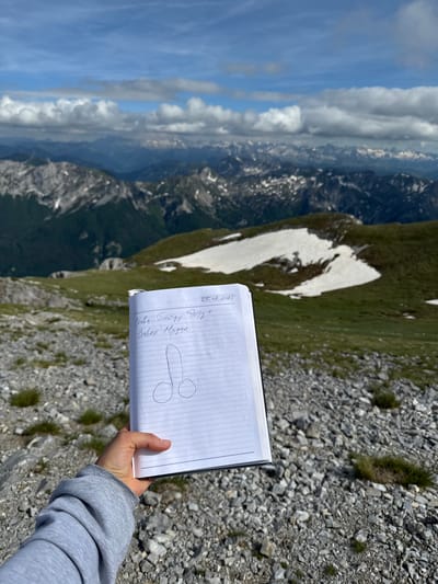 An outdoor, medium shot shows a notebook held in a person's left hand. The notebook is open to a page with handwritten text and a drawing. The text is in Cyrillic script and reads: "Слава Господу Богу + СЛАВКО Мрцик". Above the handwritten text is a date: "27.07.2021". Below the text is a drawing of what appears to be a penis. The notebook is positioned in front of a mountainous landscape under a cloudy blue sky. The mountains are covered in green vegetation and patches of snow. The ground in the foreground is rocky and dry with sparse green grass. The scene appears to be in Jošanica, Montenegro, based on contextual information.