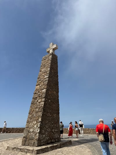 This image shows the Cabo da Roca monument, located in Colares, Portugal, which is the westernmost point of mainland Europe. A tall, stone-constructed monument topped with a cross stands prominently in the center of the frame. The monument is surrounded by a paved area and a low stone wall. Several tourists are visible, observing the monument and the sea beyond. The sky is clear and blue with some wispy clouds, and the ocean is visible in the background.