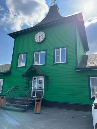 The photograph captures the facade of a vibrant green building under a bright, sunny sky with scattered clouds. The building features a prominent large clock on its upper level, just below a dark, steeply pitched roof topped with a weather vane. Several windows with white frames and dark trim are visible on the green siding. A dark canopy with ornate metalwork extends over the double glass doors leading into the building. To the left of the entrance, a wooden staircase with dark metal railings ascends to the doorway. Two wooden trash receptacles are positioned on either side of the staircase. The ground in front of the building is paved. On the right side of the image, a portion of a white car is visible. The overall scene appears to be an exterior view of a commercial or public building.