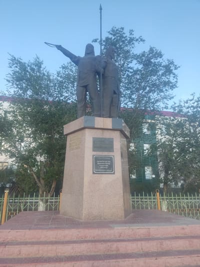 The image shows a bronze monument of two men on a stone pedestal in the city of Aral, Kazakhstan. The monument is located outdoors with trees and a building in the background. The man on the left is pointing with his right hand, holding a chain-like object. The man on the right is holding a spear. The pedestal has inscriptions on it. The monument is surrounded by a metal fence and is accessed by stone stairs.