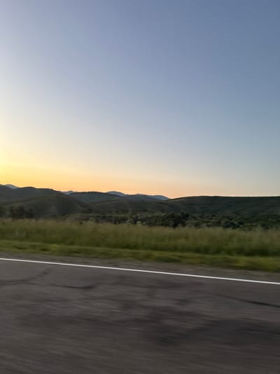 The image shows a wide, panoramic view of a rural landscape with rolling hills under a clear, gradient sky that transitions from pale yellow near the horizon to a soft blue overhead. The sun is low, casting a warm glow on the distant hills, which are covered in dark green vegetation. In the foreground, a paved road with a white line marking the lane stretches across the frame, showing motion blur suggesting it was taken from a moving vehicle. To the left of the road, a strip of blurred green grass and vegetation runs along the roadside. The overall impression is one of a serene, undeveloped natural environment. This scene is consistent with the rural surroundings of Severnoe, Kazakhstan, which features open landscapes and agricultural land.