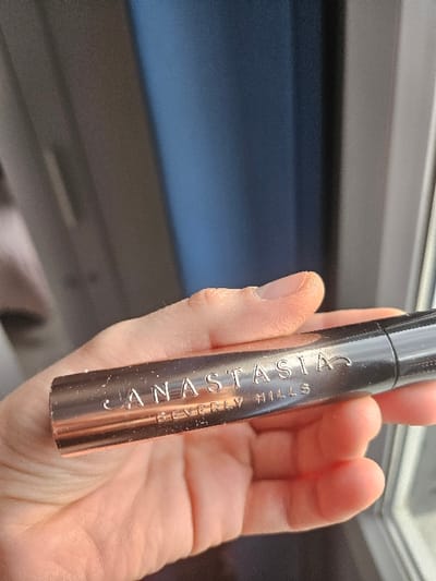 The scene is located in Villeneuve-Saint-Georges, France. An adult's hand holds a rose gold-colored cosmetic product horizontally in a close-up, upward-angled shot. The cylindrical product features the embossed text "ANASTASIA" prominently, with "BEVERLY HILLS" inscribed beneath it. The metallic surface of the product exhibits minor abrasions. One end of the product is dark, possibly black, suggesting a cap or applicator. The background is blurred, showing a light-toned area to the left and a dark blue-grey rectangular element to the right, consistent with an indoor environment receiving natural light, likely from a window.