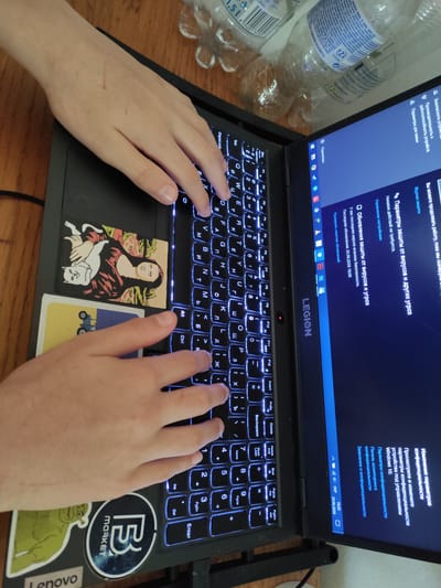 The image shows a close-up, overhead view of a person's hands typing on a laptop. The laptop is a dark color with illuminated keys, and the screen is displaying text in Russian related to computer security settings. There are stickers on the laptop, including one with a depiction of the Mona Lisa with a cat, and another with a green cartoon character. The background shows a wooden surface and parts of plastic bottles. The overall scene appears to be indoors, possibly a desk setting. The image does not contain any identifiable landmarks or elements that would indicate a specific location in Jablonec nad Nisou, Czechia.