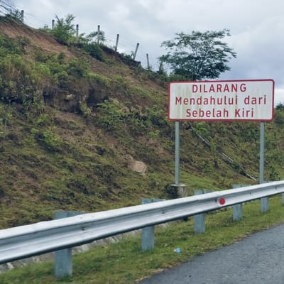 The image captures a roadside scene in Seulimeum, Indonesia, under an overcast sky. In the foreground, a silver-colored metal guardrail stretches across the lower part of the frame, alongside a dark asphalt road, of which only a small portion is visible at the bottom right. A red circular reflector is affixed to the guardrail, and a small blue plastic bottle lies discarded on the grass below it.

To the right, a prominent rectangular white sign with a red border stands on two metal posts. The sign displays text in red, in Malay/Indonesian:
"DILARANG
Mendahului dari
Sebelah Kiri"
This translates to "NO OVERTAKING from the Left Side."

Behind the guardrail and the sign, a steep, rugged hillside rises, covered in a mix of earthy brown soil and varying shades of green vegetation, including grass, shrubs, and some scattered trees. Signs of erosion are visible on the slope. Along the crest of the hill, a simple fence with concrete posts and barbed wire is noticeable. The sky above is uniformly grey and cloudy, suggesting an overcast day. No people or moving vehicles are visible, contributing to a serene yet regulatory roadside atmosphere.