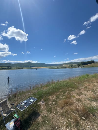 The image shows a wide, tranquil lake with a backdrop of rolling, green hills under a bright blue sky dotted with fluffy white clouds. A prominent sun glare streaks across the upper left quadrant of the sky. The foreground features a grassy, uneven terrain sloping down towards the water's edge. On the left, a foldable fishing chair is set up next to a small, portable table. The table is laden with fishing equipment, including fishing rods propped up nearby, what appears to be tackle boxes, a book, and some packaged items. A cooler and a bag are positioned on the ground near the chair. In the water, two dead, skeletal trees rise eerily from the surface, adding a stark contrast to the otherwise serene landscape. The scene depicts a peaceful, outdoor setting, likely a popular fishing spot. The provided metadata indicates the location is Drenkovo, Bulgaria.