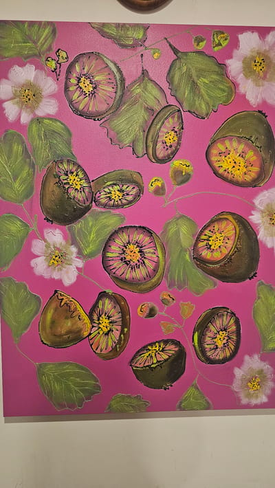 This is a close-up, head-on photograph of a painting of kiwifruit and flowers on a bright pink background. The painting features several whole kiwifruit, sliced kiwifruit, green leaves, and delicate white and pink flowers. The kiwifruit have a textured brown exterior and a vibrant, star-shaped interior with yellow seeds. The leaves are painted in various shades of green with outlines in black. The flowers have multiple petals and yellow centers. The overall style is somewhat stylized and decorative. The painting is displayed on a wall, and the top edge of a wooden frame is visible at the very top of the image. The bottom of the painting is at the bottom edge of the frame, and a white wall is visible below it. This image does not contain information about the city of Henderson-Massey, New Zealand.
