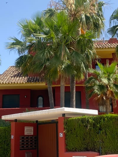 This media file shows a red building with a yellow trim, partially obscured by lush green palm trees. The building has a tiled roof and several windows. A white awning extends from the building, and a neat green hedge lines the side. The scene appears to be in a sunny outdoor location, possibly a resort or residential complex, given the tropical vegetation and well-maintained appearance. The sky is a clear blue, suggesting good weather.
Visible text includes the number "17" and "15" in blue squares, likely indicating apartment or unit numbers. There is also a small white sign on the building with Spanish text that reads: "AVISO PARA VISITANTES / PUBLICIDAD: 'Todo publicidad en este mismo edificio, debe tener anuncios de particulares, queda prohibida la empresa anunciante. LA COMUNIDAD DE VECINOS'". This translates to "NOTICE FOR VISITORS / ADVERTISING: 'All advertising in this building must have private advertisements, it is forbidden by the advertising company. THE COMMUNITY OF NEIGHBORS'". The time of day appears to be daytime, given the bright sunlight and shadows. The image has a vertical orientation, and the palm trees are swaying, indicating a gentle breeze. There are no people visible in the media file.